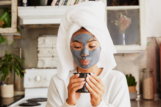 Woman with clay face mask and towel wrapped around head, enjoying a cup of tea in her cozy kitchen.