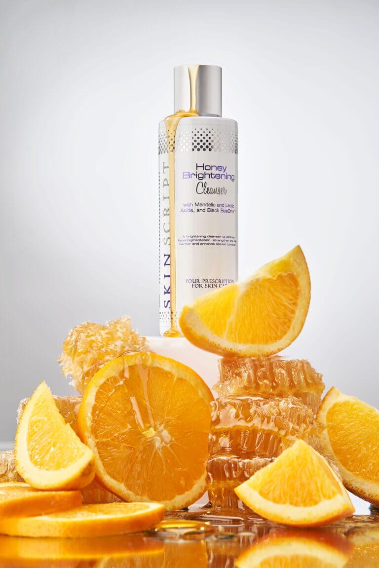 Honey Brightening Cleanser with 2.5% mandelic and lactic acid, surrounded by honeycomb and fresh orange slices for brightening benefits.