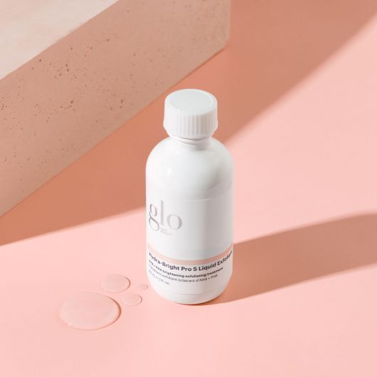 Hydra-Bright Pro 5 Liquid Exfoliant bottle on a pink surface with drops of liquid, designed to smooth, refine, and brighten skin.